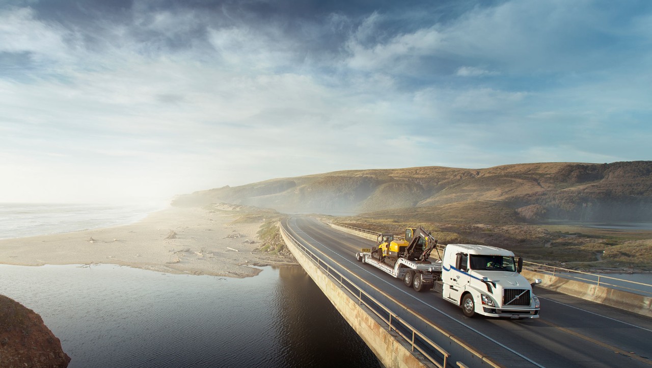 White Volvo truck on a bridge carrying a yellow excavator